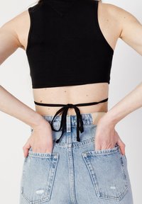 Kobieta w czarnym topie na krótki rękaw z zawiązanym tyłem oraz w jasnoniebieskich dżinsach o wysokim stanie, z rękami w tylnych kieszeniach.