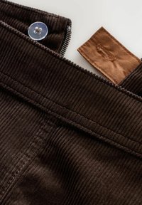 Tessuto in corduroy marrone con righe verticali, caratterizzato da una chiusura a bottone e un'etichetta in tessuto marrone, posizionata sulla vita.