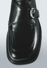 Zwarte leren schoen met een afgeronde neus, een gekruiste band met een gesp en ingewikkeld stiksel langs de rand. Gladde, glanzende oppervlaktestructuur.