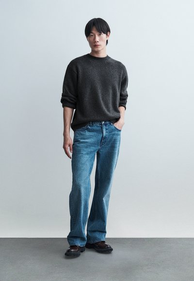 Jeune homme debout à l'intérieur portant un pull gris foncé, un jean bleu clair et des chaussures marron, sur un fond uni gris clair.