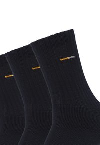 Sorte ribbede bomuldsokker med elastisk top, der har et lille orange og hvidt logo nær manchetterne. Tre par er vist.