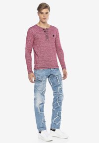 Rotes Langarm-Henley-Shirt mit geknöpftem Ausschnitt und Logo, kombiniert mit hellblauen, ausgewaschenen Jeans und weißen Sneakern.
