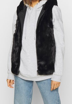 Personne portant un sweat à capuche gris clair et un gilet en fausse fourrure noire, associé à un jean bleu clair, visible du cou aux hanches.