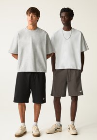 Dva modely nosia voľné oblečenie: šedé a biele tričká s oversized strihom v kombinácii s čiernymi a šedými šortkami, ktoré majú jednoduché vzory a akcenty loga.