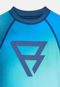 Rash guard z modrim gradientom, izdelan iz sintetičnega materiala. Ima temno modro logo in kontrastno šivanje vzdolž šivov. Gladka tekstura.