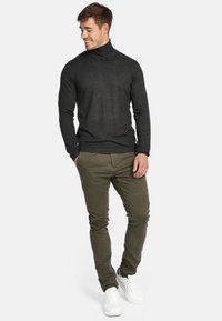 Mørk grå genser med høy hals, tettsittende design, kombinert med olivengrønne slim-fit bukser. Hvite joggesko fullfører antrekket.
