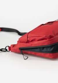 Borsa in tessuto impermeabile rosso con una superficie liscia, dotata di una cerniera nera e dettagli in metallo, forma ergonomica e tracolla regolabile.