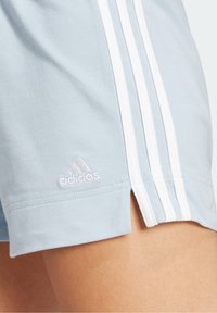 Helleblaue Adidas-Shorts mit weißen drei Streifen und Logo-Stickerei auf dem Oberschenkel einer Person.