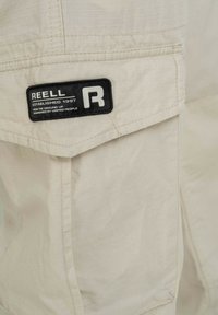 Beige cargoshorts gemaakt van lichtgewicht stof met een klein zwart label met witte tekst. Horizontale stiksels zijn zichtbaar.