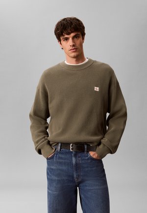 Junger Mann mit Händen in den Taschen trägt einen olivgrünen Strickpullover und blaue Jeans vor einem einfarbigen grauen Hintergrund.