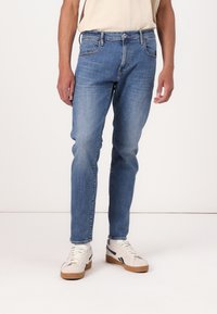 Jeans de denim albastri estompați cu o croială dreaptă, cinci buzunare și închidere cu un buton metalic, asortați cu pantofi sport albi și bej.