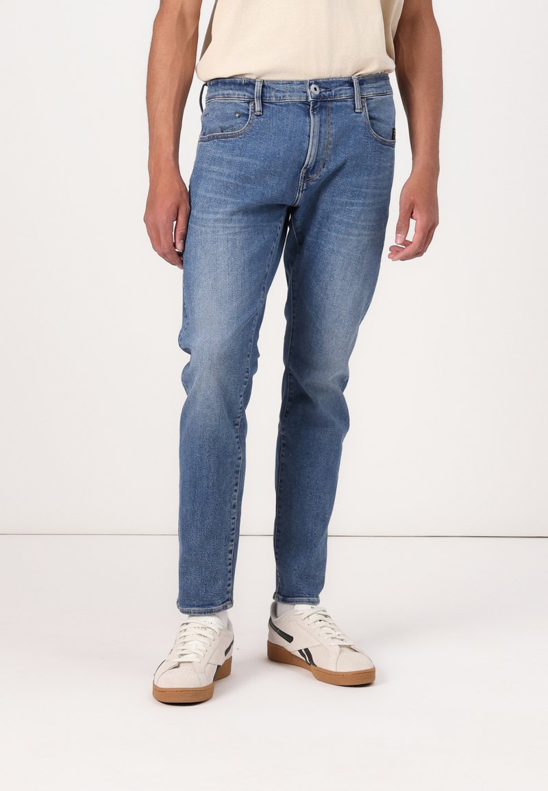 Jeans de denim albastri estompați cu o croială dreaptă, cinci buzunare și închidere cu un buton metalic, asortați cu pantofi sport albi și bej.