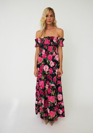 Frau mit langen blonden Haaren, die ein schwarzes schulterfreies Maxikleid mit knalligen pinken und roten Blumenmustern trägt, steht vor einem schlichten hellen Hintergrund.