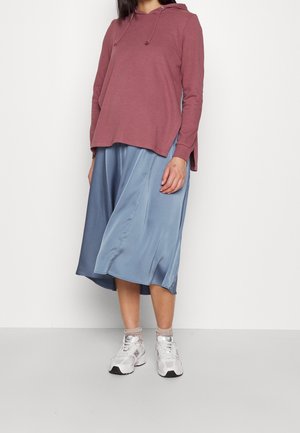 Personne portant un sweat à capuche rose poudré, une jupe midi en satin bleu, des chaussettes beige et des baskets blanches, debout devant un fond blanc uni.