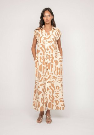 Une femme se tient debout portant une robe maxi sans manches à motifs beige et marron clair avec des poches et des sandales beige à lanières.