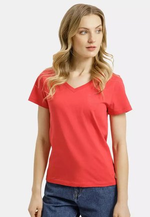 Kobieta o długich, falistych blond włosach, ubrana w czerwony t-shirt z krótkim rękawem i dekoltem w serek oraz niebieskie dżinsy, patrząca lekko w prawo.