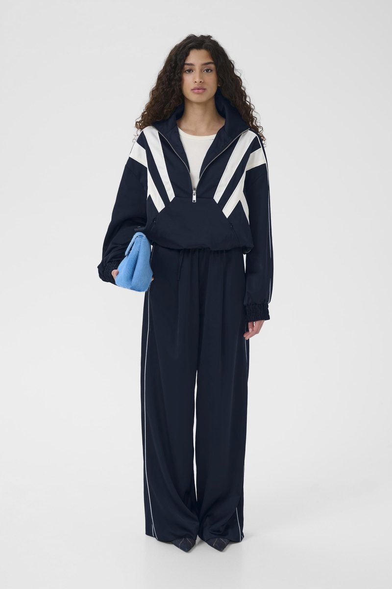 Marineblauer Trainingsanzug mit einer Reißverschlussjacke mit weißen diagonalen Streifen und passenden lockeren Hosen. Das Modell hält eine blaue Tasche.