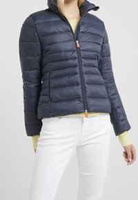 Doudoune marine avec une fermeture éclair sur le devant, un motif matelassé horizontal, un col montant et un tire-zip orange contrastant. Présente une coupe cintrée.