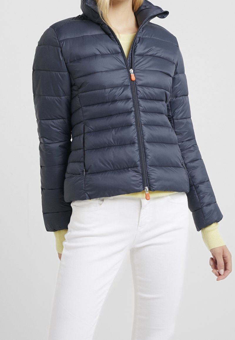 Doudoune marine avec une fermeture éclair sur le devant, un motif matelassé horizontal, un col montant et un tire-zip orange contrastant. Présente une coupe cintrée.