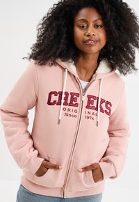 Felpa con zip di colore rosa chiaro, foderata in finto pelo bianco, con logo rosso ricamato, cappuccio con lacci e tasche frontali. Tessuto morbido e vestibilità rilassata.
