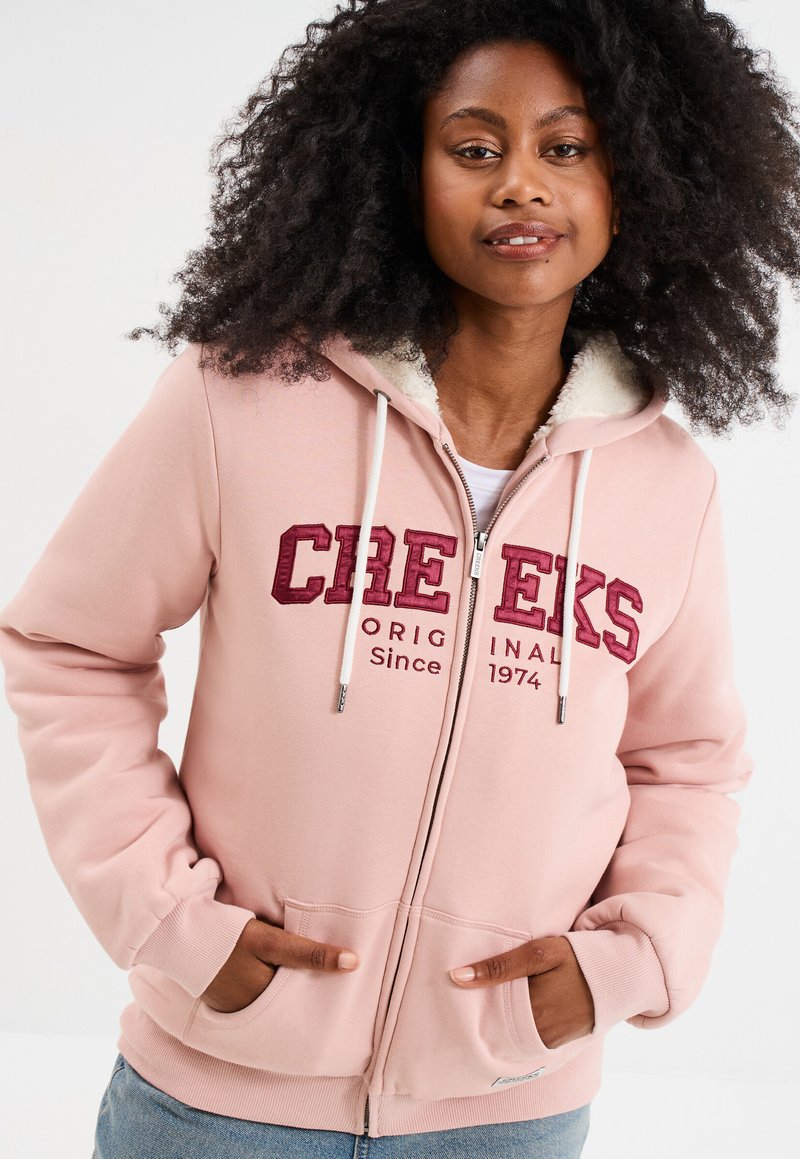 Felpa con zip di colore rosa chiaro, foderata in finto pelo bianco, con logo rosso ricamato, cappuccio con lacci e tasche frontali. Tessuto morbido e vestibilità rilassata.