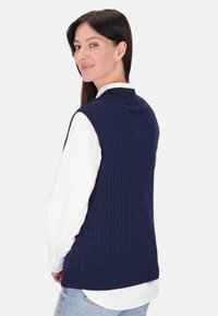 Námořnická pletená vesta s ribbed texturou a kulatým výstřihem, navlečená přes bílou košili, která ukazuje hladkou a úzkou siluetu.