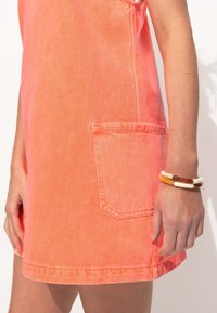 Robe en denim corail avec une poche latérale, présentant une coupe décontractée et des coutures visibles. Portée avec un bracelet multicolore au poignet.