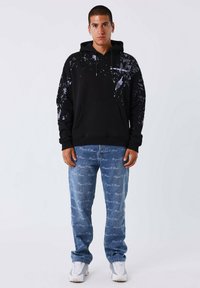 Felpa nera con design schizzitoso in viola, tasca a marsupio e coulisse regolabile. Abbinata a jeans in denim azzurro chiaro con testo ripetuto.