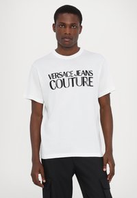 Biała bawełniana koszulka z wyrazistym czarnym nadrukiem "VERSACE JEANS COUTURE", okrągłym dekoltem i krótkimi rękawami.