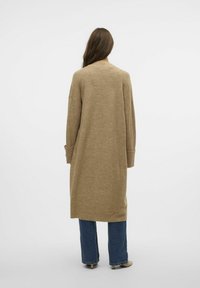 Langer beiger Cardigan mit lockerer Passform, überschnittenen Schultern und gerippten Bündchen, kombiniert mit hochgeschnittenen blauen Jeans und neutralen Schuhen.