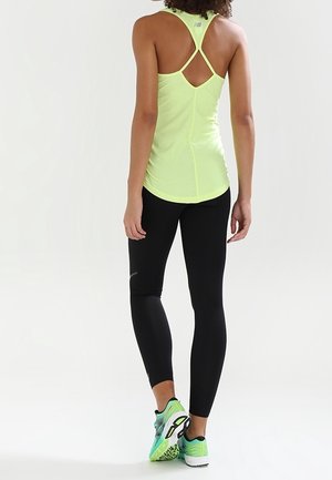 Vrouw staat met haar rug naar de kijker, draagt een gele racerback tanktop, zwarte leggings en groene hardloopschoenen op een witte achtergrond.