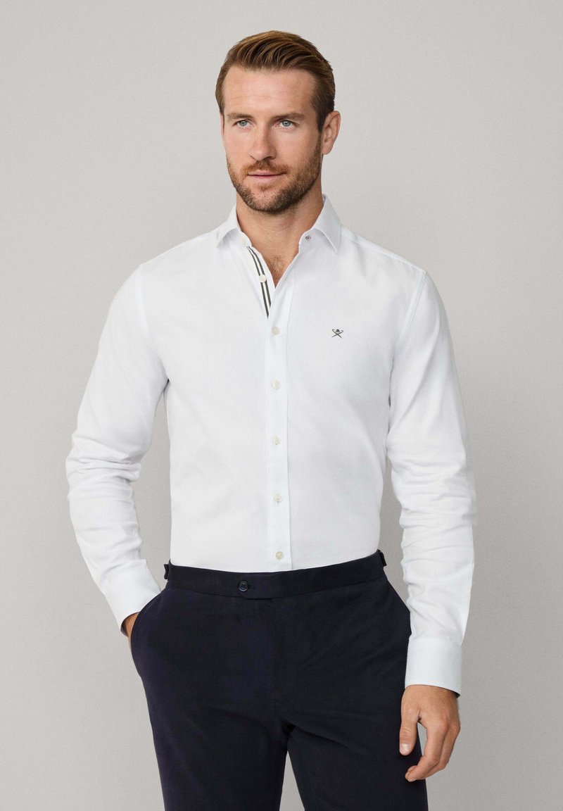 Chemise blanche à col classique, à manches longues, avec un logo discret sur la poitrine ; assortie à un pantalon bleu marine foncé.