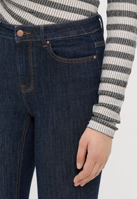 Μαύρο τζιν παντελόνι σε σκούρο μπλε χρώμα με αντίθετη πορτοκαλί ραφή, σχέδιο πέντε τσεπών και κουμπί στη μέση. Ορατός ριγέ μανίκι σε μαύρο και λευκό.