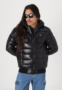 Zwarte gewatteerde jas met een gestikt patroon en een ritsvoorkant, met een capuchon en logo-accenten. Draag het met een grijs shirt en blauwe jeans.