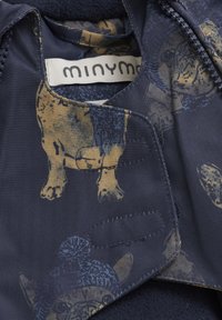 Mörkblått tyg med fiskbensmönster, prytt med ett lekfullt djurmotiv i guld- och blåtoner. Etiketten säger "minym". Mjuk textur.