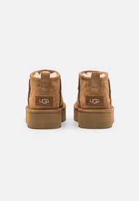 Stivaletti in suede marrone castano con una morbida fodera in pelliccia, suola piatta in gomma e tiranti sul retro. Dettaglio logo UGG sul tallone.
