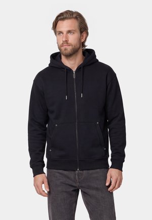Man with beard wearing black zip-up hoodie with hood up and dark gray jeans standing against light background.