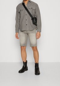 Camicia grigia con bottoni e due taschini sul petto, pantaloni corti di denim grigio chiaro e stivali neri alla caviglia. Borsa a tracolla nera indossata sulla spalla.
