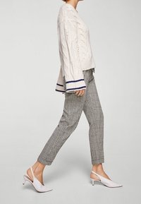 Gruby sweter w warkoczowy splot w kremowym kolorze z granatowymi mankietami; szare, tapered spodnie w kratę; białe, zapięte na pięcie buty na niskim obcasie.