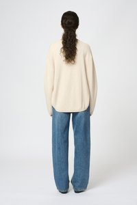 Beige oversized tröja med rundad nederkant, kombinerad med löst sittande blå denimjeans. Mjuk textur synlig på tröjan.