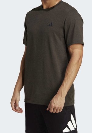 Homme portant un t-shirt Adidas vert olive foncé et un short noir avec logo Adidas blanc, debout devant un fond clair uni.