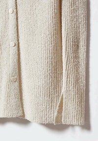 Cardigan couleur crème avec texture côtelée, comportant cinq boutons, des fentes latérales et un tissu tissé doux.