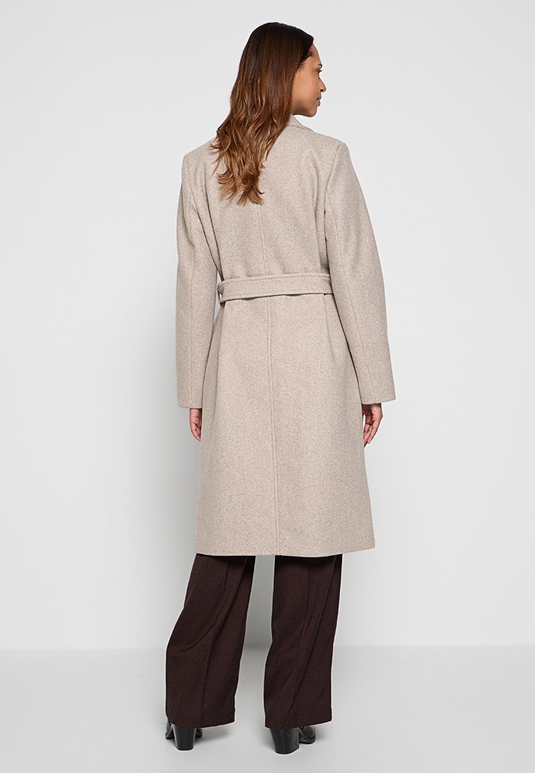 Frau steht mit dem Rücken zur Kamera, trägt einen beigefarbenen, taillierten langen Mantel, dunkelbraune weite Hose und schwarze High Heels vor einem einfarbigen Hintergrund.