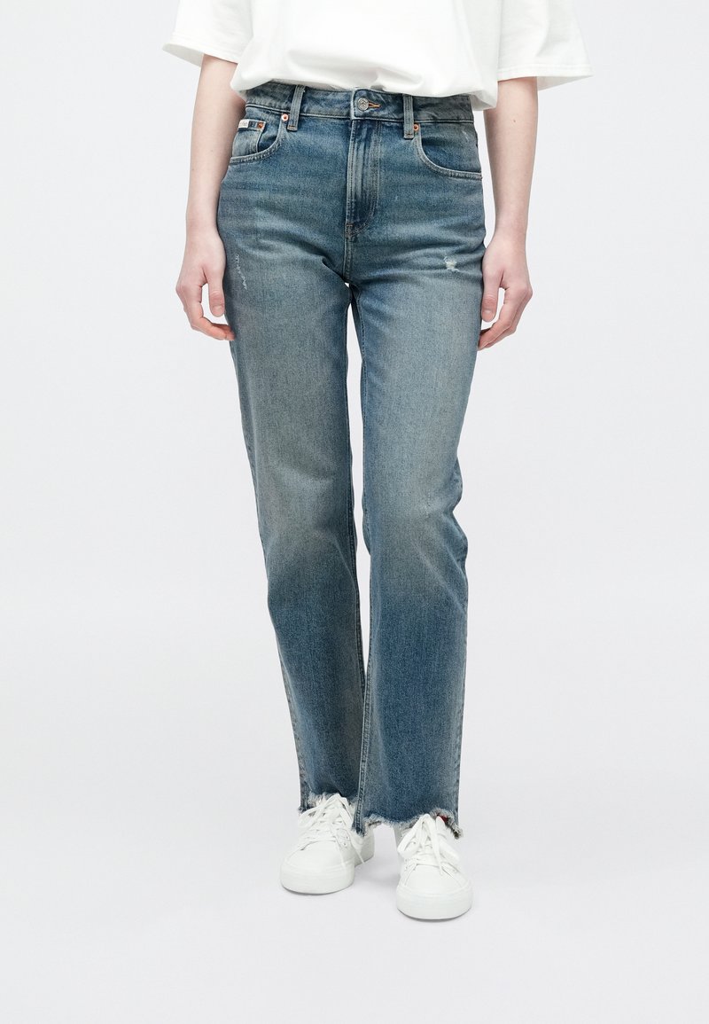 Jeans de mezclilla azul claro con un corte de pierna recta, cintura alta, detalles desgastados, dobladillo deshilachado, aspecto envejecido, combinados con zapatillas blancas.