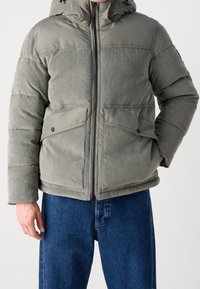 Szara kurtka puchowa z sztruksu z kapturem, z zamkiem błyskawicznym z przodu, dwoma przednimi kieszeniami oraz prążkowaną fakturą. Noszona razem z niebieskimi jeansami.