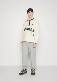 Kremfarget fleecegenser med svart "DOUBLE A" tekst, grå treningsbukser og svarte og gule joggesko. Modellen har på seg en svart caps.