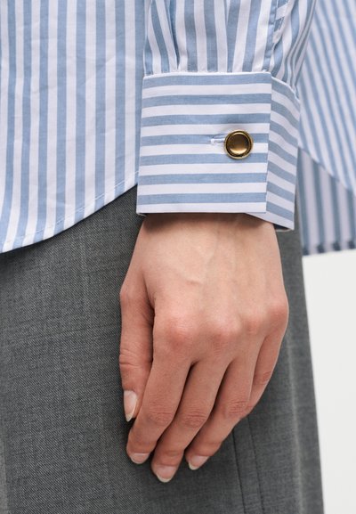 Χέρι που ακουμπά στο πλευρό, φοράει μπλε και λευκό ριγέ πουκάμισο μακρυμάνικο με χρυσό μανικετόκουμπο και γκρι παντελόνι.