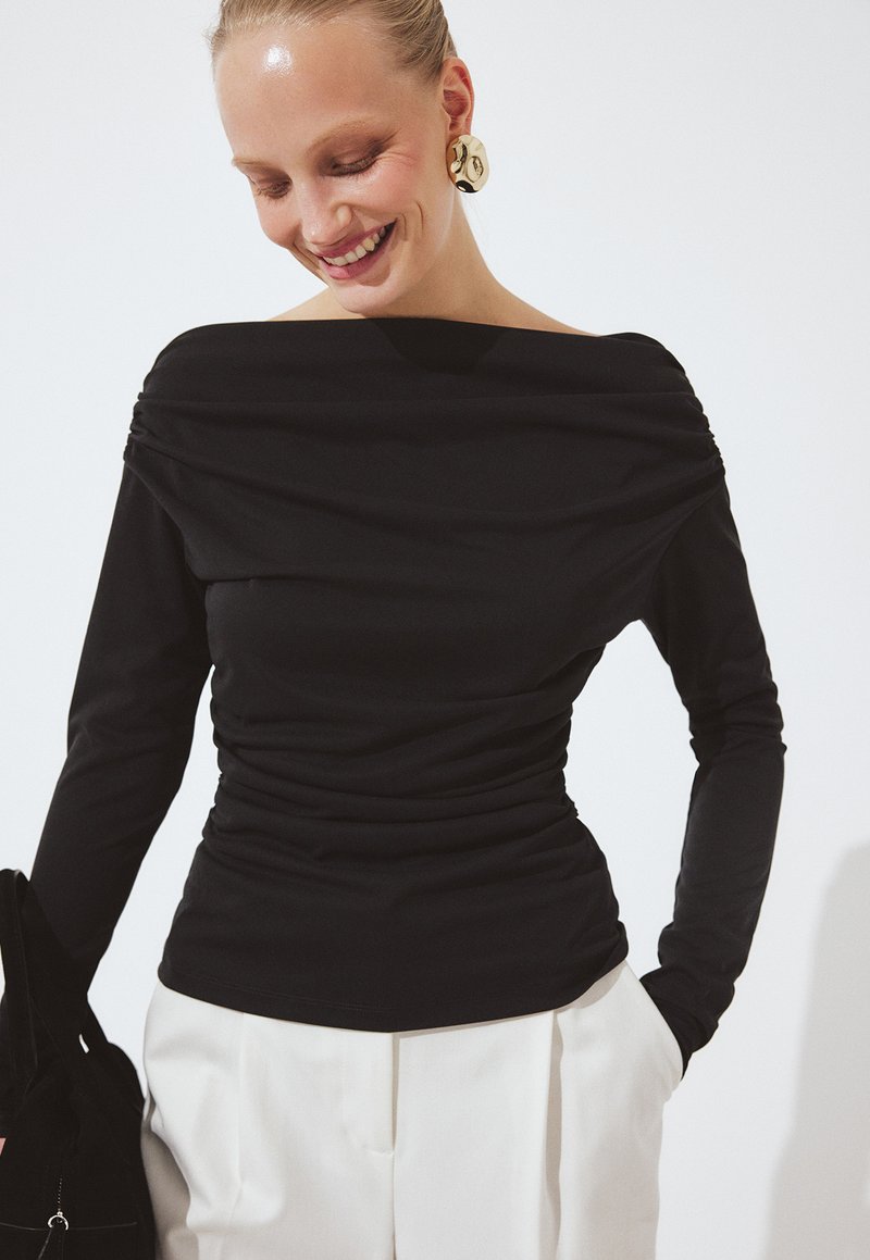 Ženska, ki se smehlja, oblečena v črno majico z dolgimi rokavi brez ramen, z nabrano stranjo, bele hlače z visokim pasom in velike zlato uhanov.