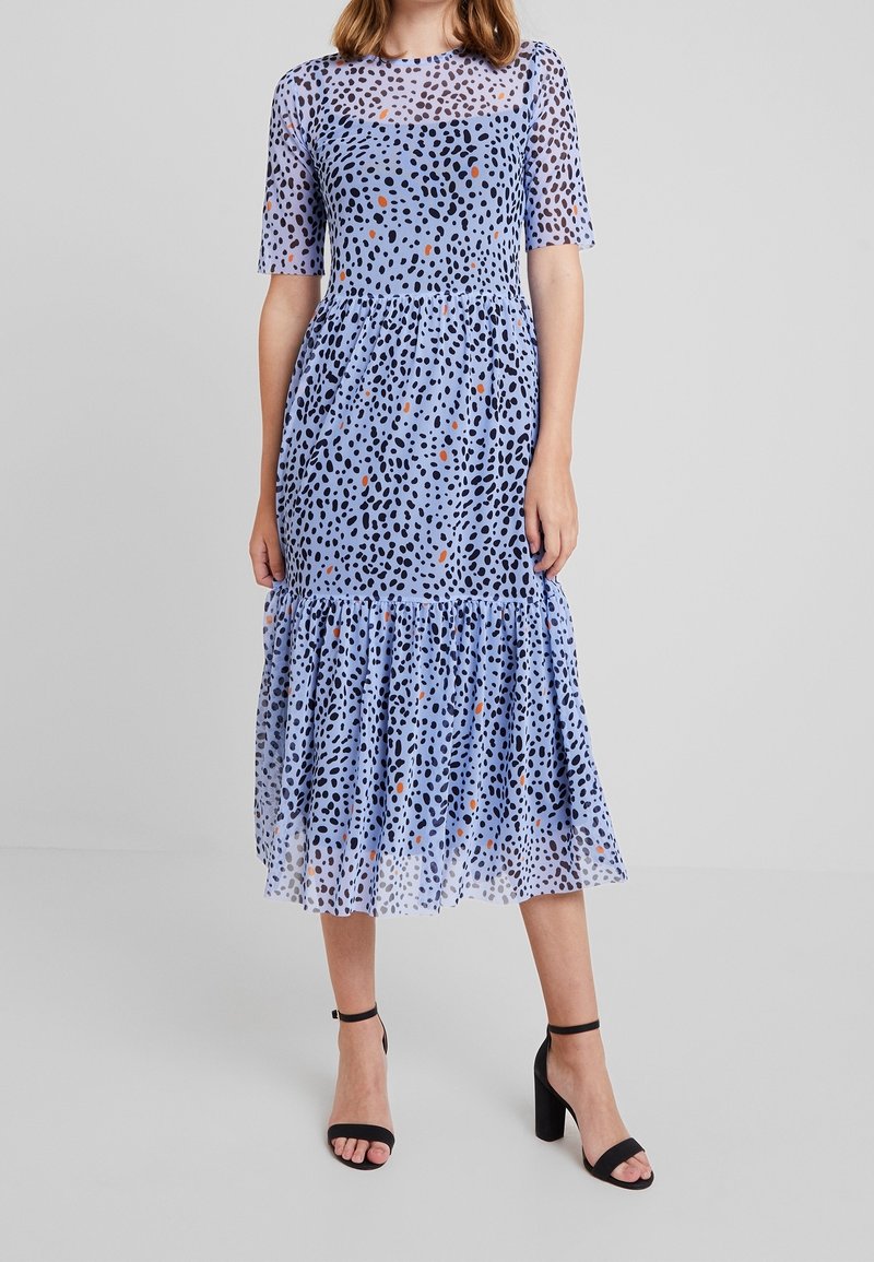 Modré šaty s černým a oranžovým skvrnitým vzorem, krátkými rukávy, přiléhavým trupem a volánkovou sukní. Nosené s černými lodičkami na kotníkovém pásku.