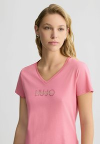 Rosa Baumwoll-T-Shirt mit V-Ausschnitt und kurzen Ärmeln. Mit einem diamantenbesetzten Logo auf der Brust. Glatte Textur und lässiges Design.